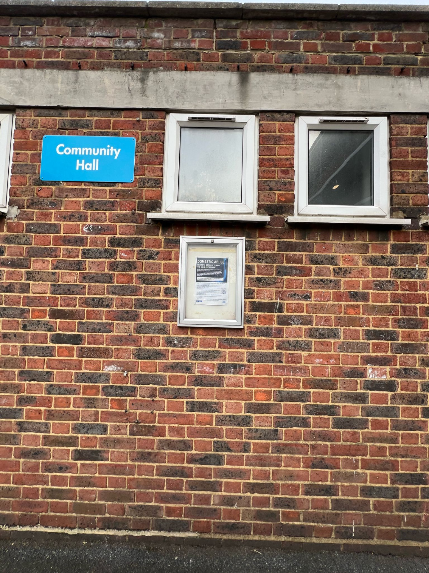 Exterior detail of The Old Workshop: a blue Community Hall sign and a glazed noticeboard on a red brick wall beneath two white-framed windows.